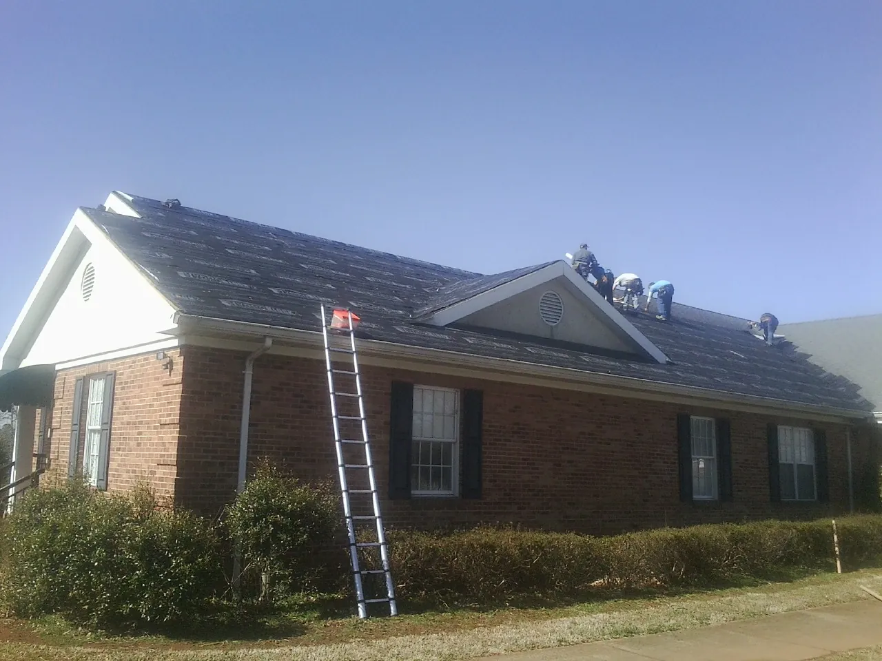 Crew tearing shingles off a brick ranch roof with a ladder set — Metal Roof Repair work by our crew in Damon