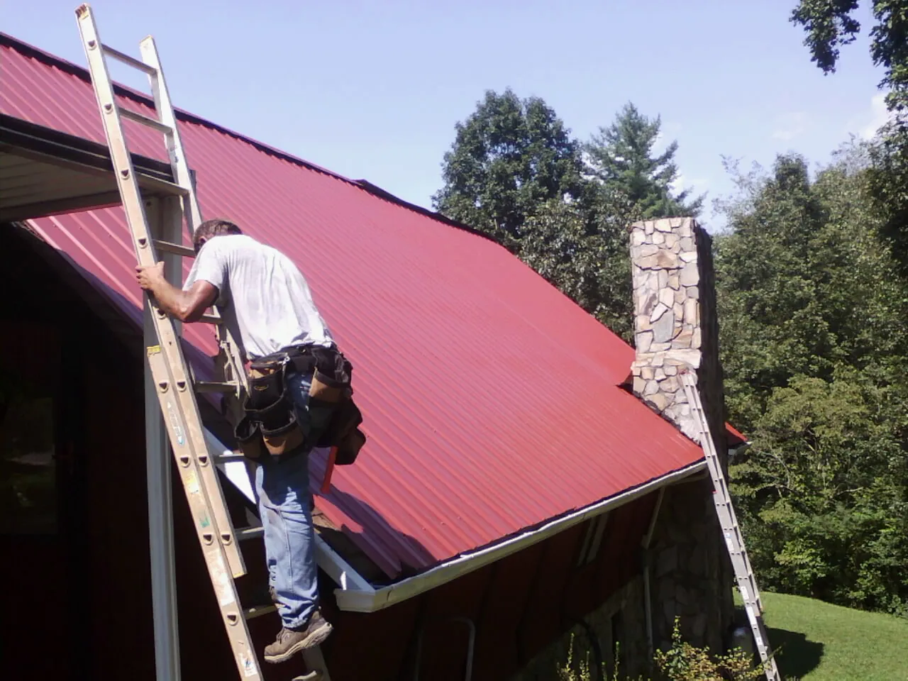 Roofer on a ladder beside a red metal roof and stone chimney — Soffit & Fascia Repair by Roofing Brothers in Damon